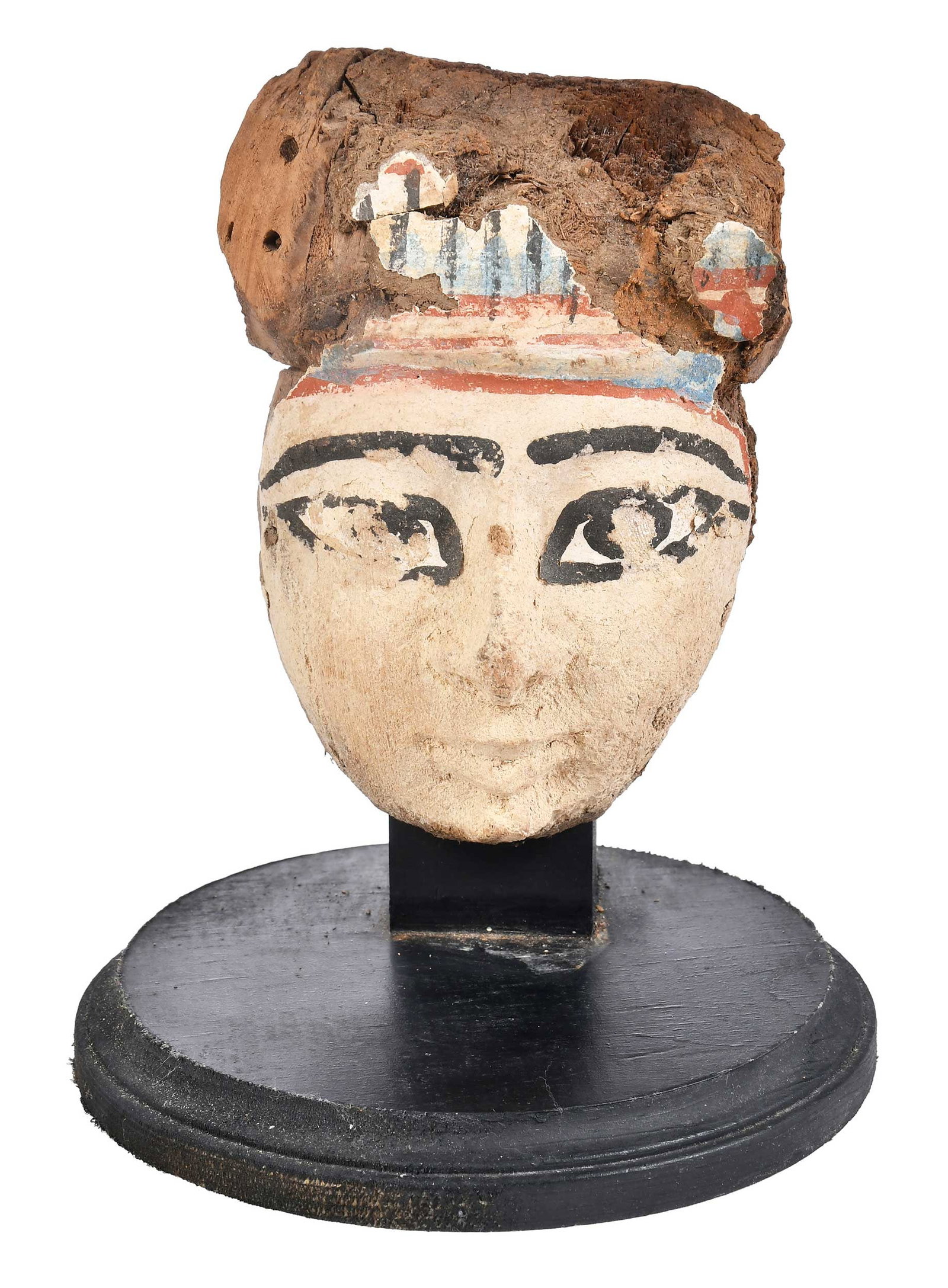 Egyptian Carved and Polychromed Cartonnage Mummy Head: possibly Late period, cartonnage on wood with white, red, blue, and black painted features, pleasant expression with wide eyes, remnants of colorful headdress, two holes with peg remnants in upper pro