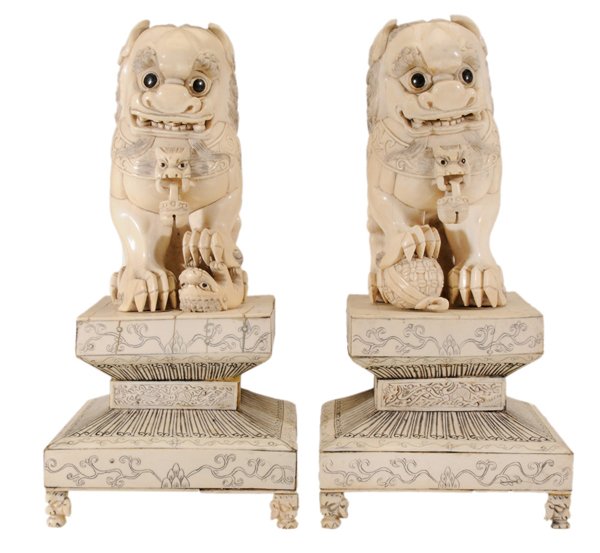 Pair Carved Ivory Seated Buddhist: Style Foo Dogs Chinese, 20th century, each with bells and black-painted eyes, one with right foot on ball, one with left foot on foo dog cub, bottom signed, laminated elaborate stepped platform bases 