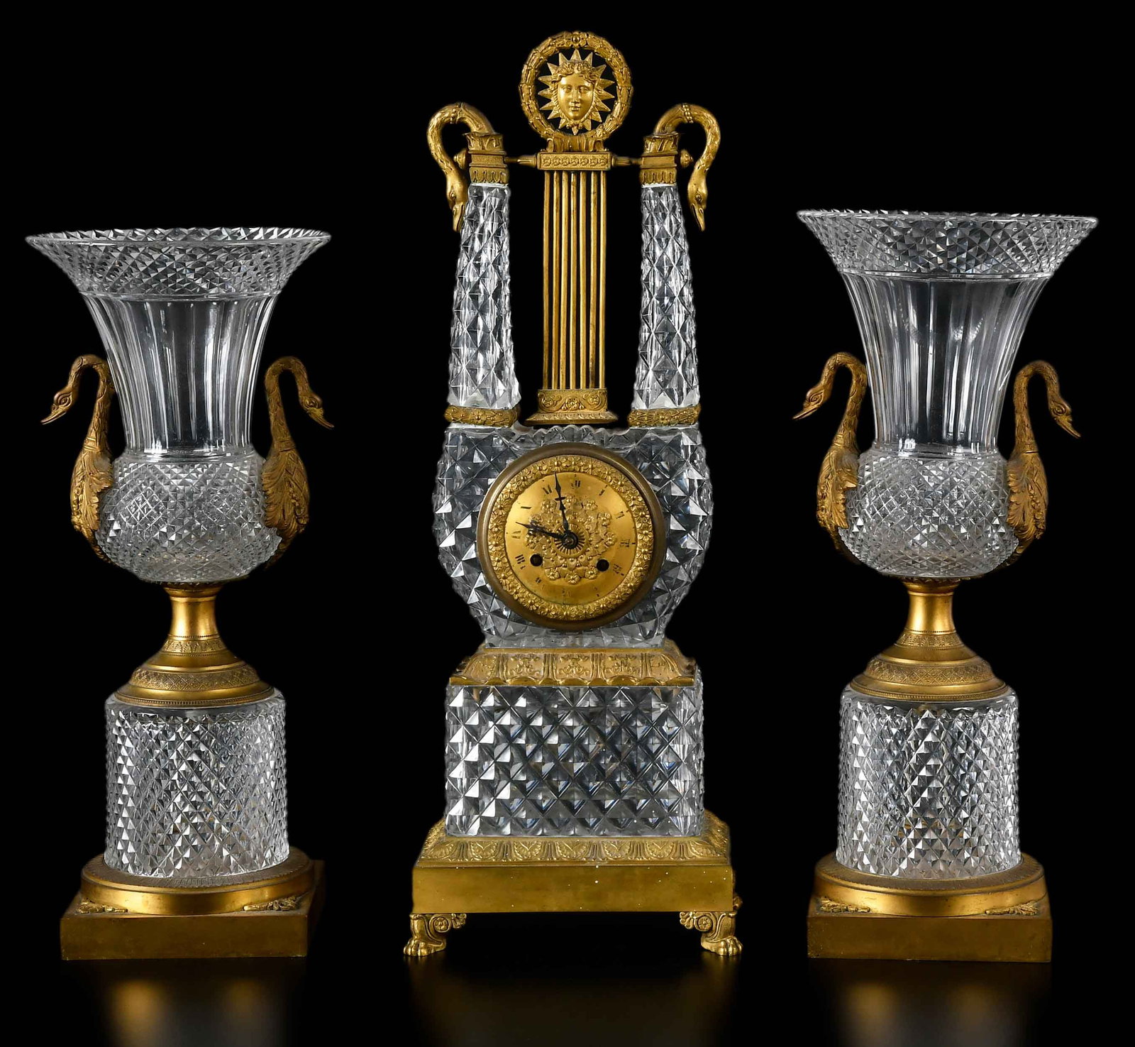French Empire or Style Three Piece Glass and Gilt Bronze Clock Garniture: late 18th/19th century, bronze dial with Roman numerals set in elaborate glass case with bronze mounts, clock 20 x 7 x 4 in.; two matching urns, 16 x 6-1/2 x 6-1/2 in. Provenance: The Collection of Jo