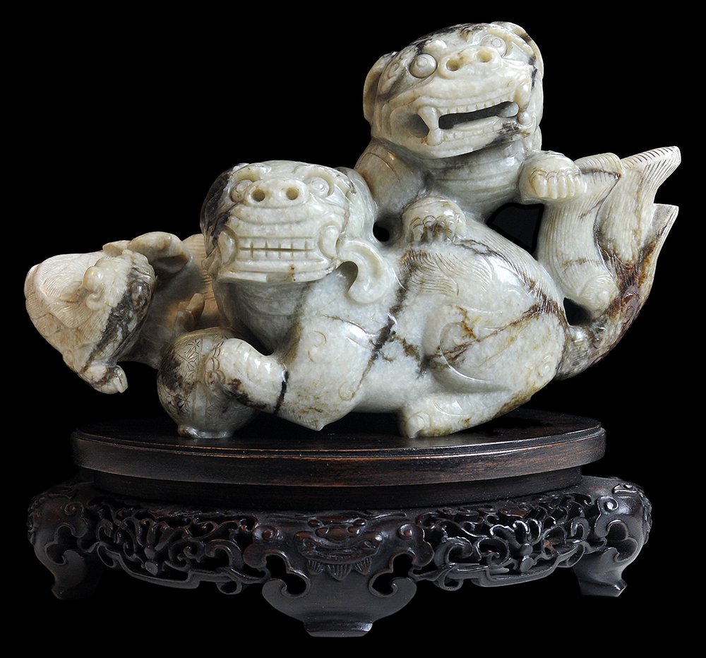 Celadon Jade Figural Group of: Buddhist Foo Dogs Playing with a Ball Chinese, 19th/20th century, playful figures depicted in celadon to pale ivory jade with russet inclusions, set on a very fine hardwood stand, 4-1/4 x 7 in., one p