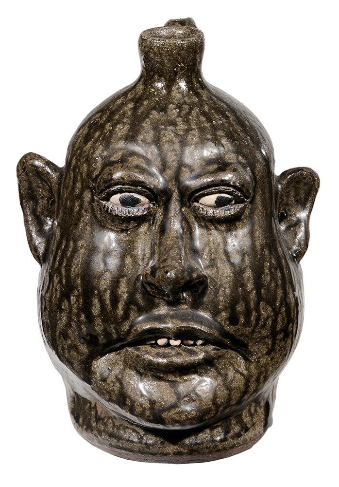 Lanier Meaders Face Jug: (White County, Georgia, 1917-1998), runny olive alkaline glaze, rock teeth, white clay eyes with painted black pupils, incised eyelashes, flared rim, applied strap handle, inscribed on base "Lanier Me