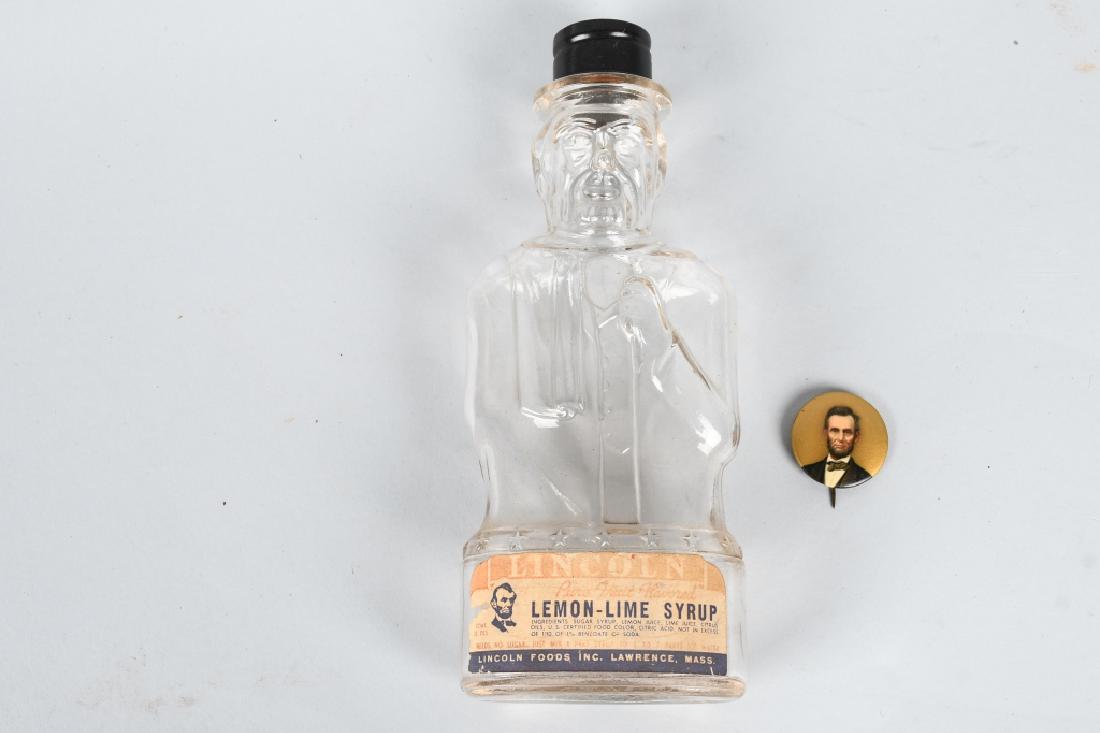 ABRAHAM LINCOLN PINBACK BUTTON & BOTTLE BANK: Abraham Lincoln pinback button measures 1 1/8" and has the original Bastian Bros paper label. Condition is Excellent. The Lincoln Syrup bottle bank has original label and litho tin cap. Very Good-Exce