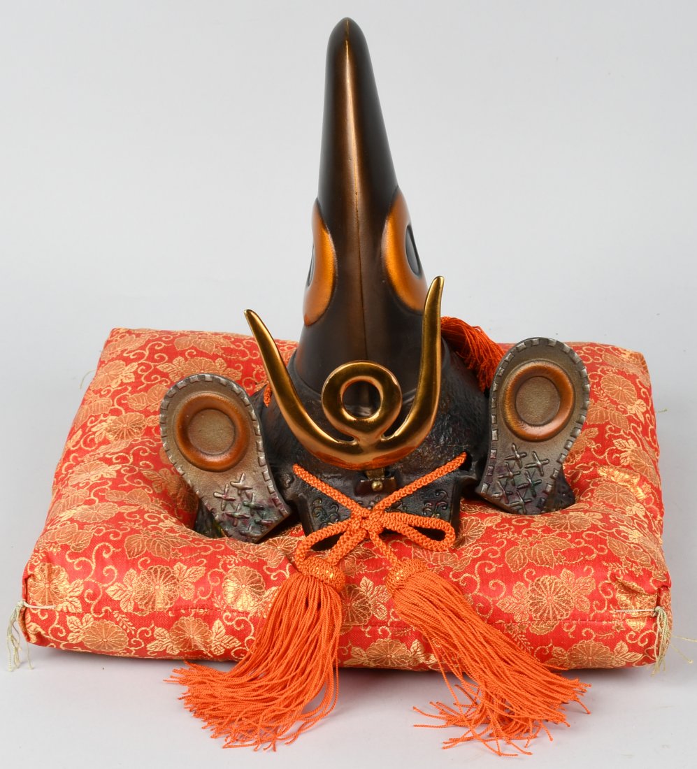 JAPANESE SAMURAI HELMET ON RESTING PILLOW: Vintage replica of an early Japanese ceremonial war helmet on brocade pillow. Made of heavy iron, with adornments and orange cords. Pillow is approximately 14.5" x 11.5" x 2" and the helmet is approxi