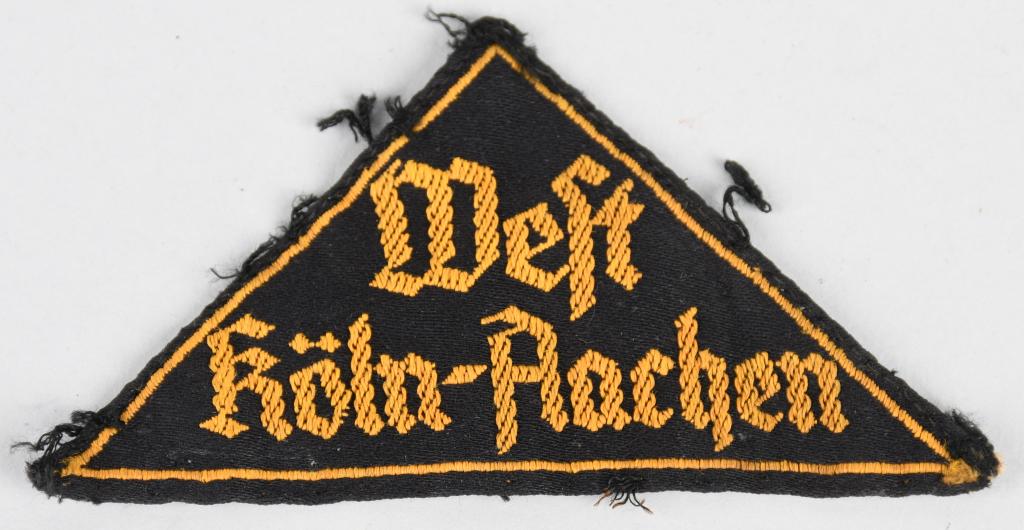 WWII HJ WEST KÖLN-AACHEN DISTRICT SLEEVE TRIANGLE: Woven black rayon construction isosceles triangle features machine woven Gothic script in two lines, "West Köln-Aachen", in golden yellow threads. The triangle also has a narrow machine woven golden 