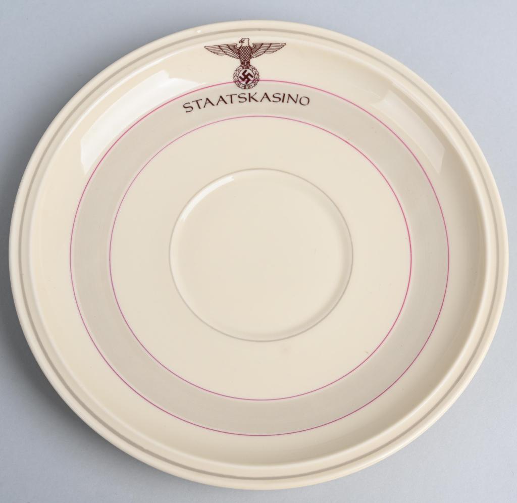 WWII NAZI GERMAN STAATSKASINO KRAKAU PLATE: Nazi German plate with red colored rings and red colored right-facing eagle over mobile swastika over the word "STAATSKASINO." Porcelain plate measuring approximately 7.5" across. Signed on the back R