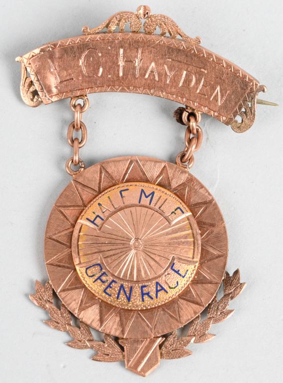 SOLID GOLD 1/2 MILE BICYCLE RACE WINNER BADGE: Named 1/2 mile bicycle race solid gold badge measuring 1 1/2 inches X 2 inches weighing 7.27 grams of gold. Top bar is named to L. C. Hayden. The drop of the badge shows a Bicycle Wheel with blue enam