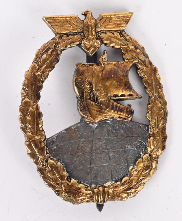 WWII NAZI GERMAN KM AUXILIARY CRUISER WAR BADGE: One piece, die struck,zinc construction badge with fire gilt and silver plated finishes. The badge is in the form a gilt washed, vertically oval, embossed, oak-leaf wreath with a national eagle with o