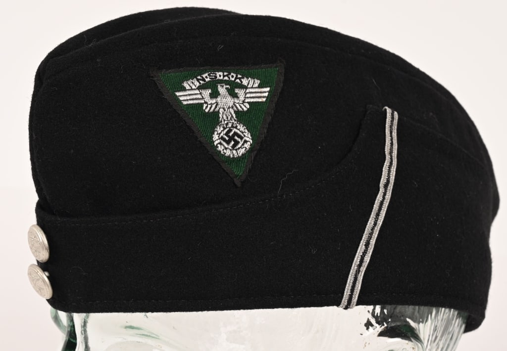 WWII NAZI GERMAN NSKK OVERSEAS CAP WW2: A black wool cap whose fold-down side panels slope towards the front where they are sewn to the side of the cap. Sewn to the left side of the cap, above where its side panel begins to slope, is a mach
