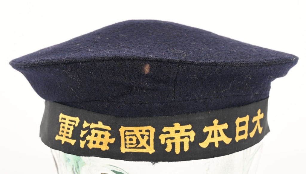 WWII IMPERIAL JAPANESE SEAMAN’S DONALD DUCK CAP: A black wool Imperial Japanese Navy seaman’s cap with a post-1941 cap tally. The cap’s 22cm diameter crown has its original stiffening wire intact under the forward seam. The headband has
