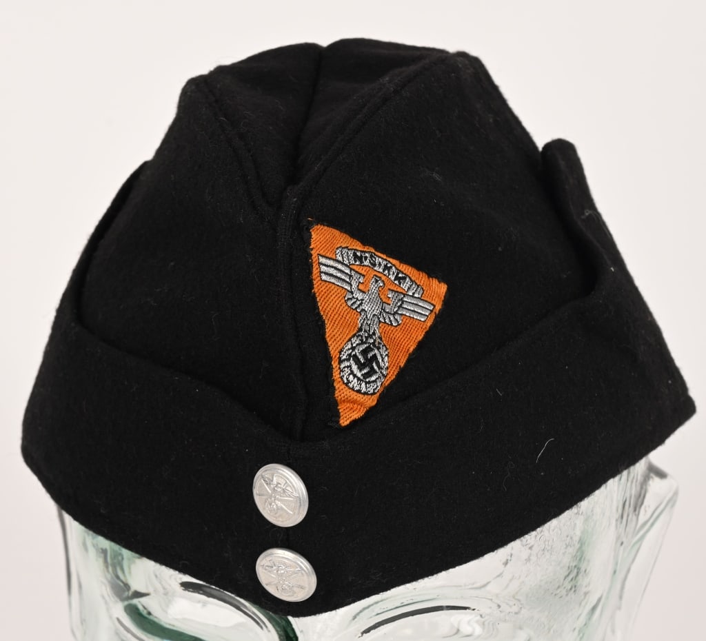 WWII NAZI GERMAN NSKK SÜDWEST OVERSEAS CAP WW2: A black wool cap whose fold-down side panels slope towards the front where they are sewn to the side of the cap. Sewn to the left side of the cap, above where its side panel begins to slope, is a mach