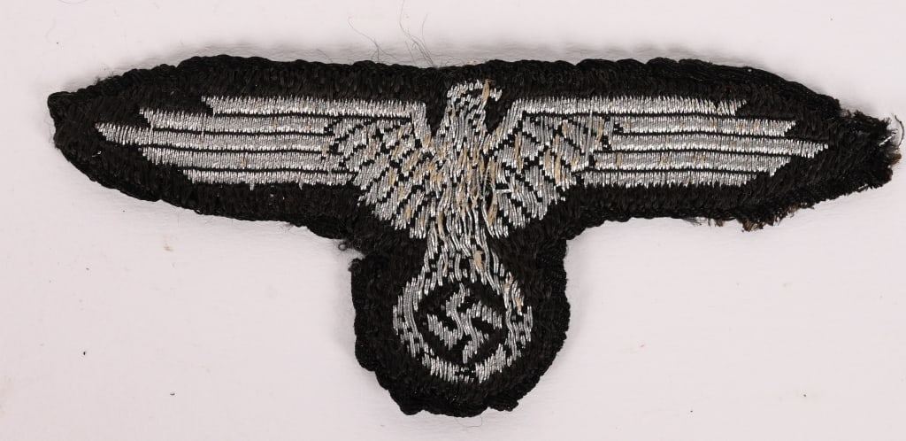 WWII NAZI SS OFFICER’S OVERSEAS M43 CAP EAGLE WW2: Machine woven, SS overseas / M43 cap eagle with out-stretched wings, clutching a wreathed, canted, swastika in its talons in silver wire threads on a cut-out, woven black rayon base. Has been removed 