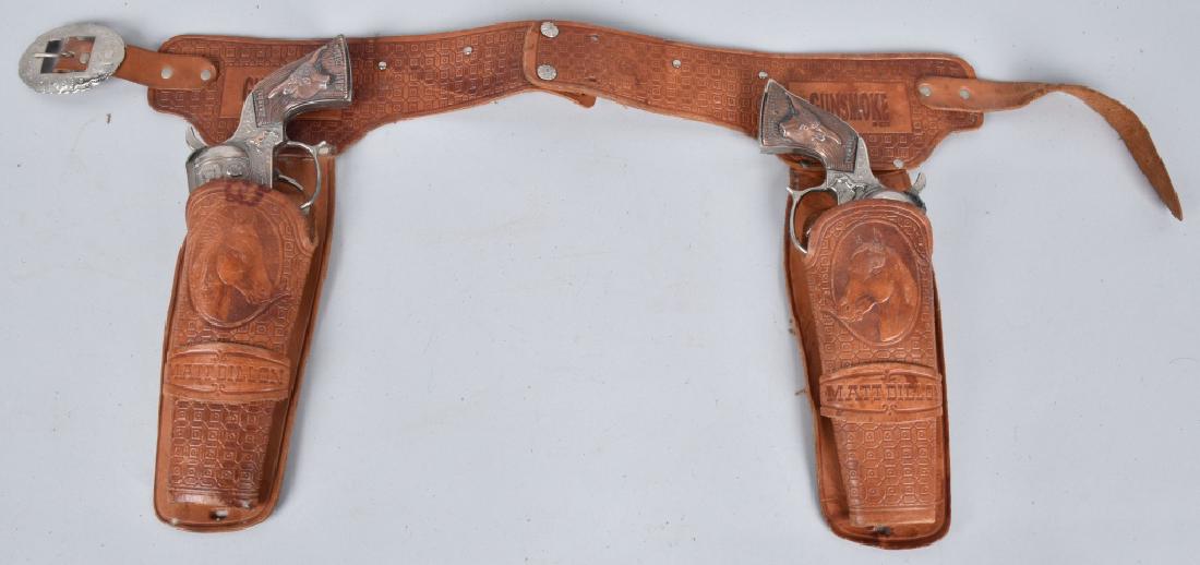 GUNSMOKE DOUBLE HOLSTER CAP GUN SET: Nice set with 2 working metal grip Gunsmoke cap pistols, along with tooled leather Gunsmoke belt and holsters. Both holster and embossed Matt Dillon. General play wear. Very Good-Excellent