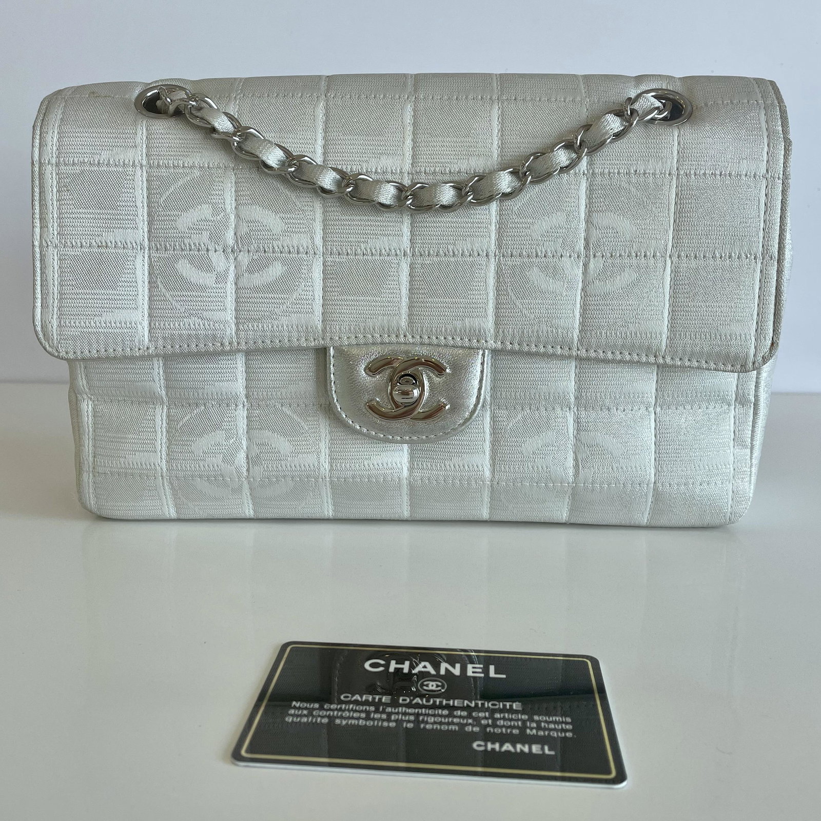 Authentic Chanel Bag Silver and White Logo Fabric Classic Flap: Guaranteed authentic. Includes authenticity card and matching hologram sticker located inside bag. Coveted medium classic flap size, measuring approx. 10"L x 2.5"W x 6.5"H. Silver toned hardware with 