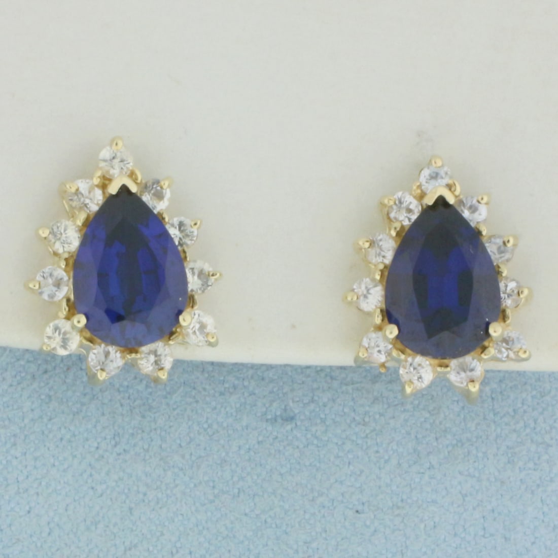 Sapphire Halo Earrings in 10k Yellow Gold: In 10k Yellow Gold. Contains two 10x7mm high quality pear cut Lab Sapphire gemstones, approximately 4ct TW, with intense color and excellent clarity. Also contains twenty-two .02ct high quality white 