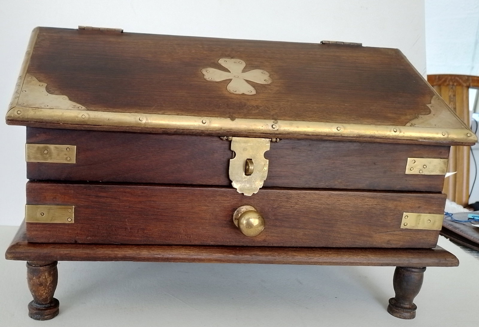 Wood and Brass Desk Tidier: Title: Wood and Brass Desk Tidier Description: This brass and wooden tidier has brass edges and a brass shamrock on the sloping lid. Stands on turned legs. There is one drawer and