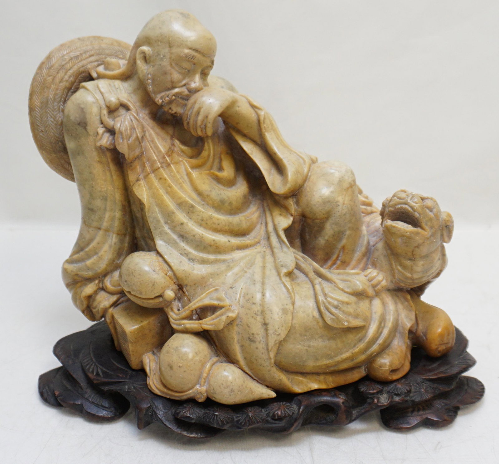 CARVED CHINESE SCHOLAR & SHISHI: Republic Era Chinese Finely Carved Soapstone Sculpture of a reclining Scholar and Shishi. On carved rosewood stand. Sculpture 8"x5"x7". Stand 8 1/2"x5 1/3"x3/4". Weight 8 pds. 4 oz. PROVENANCE: A Priv