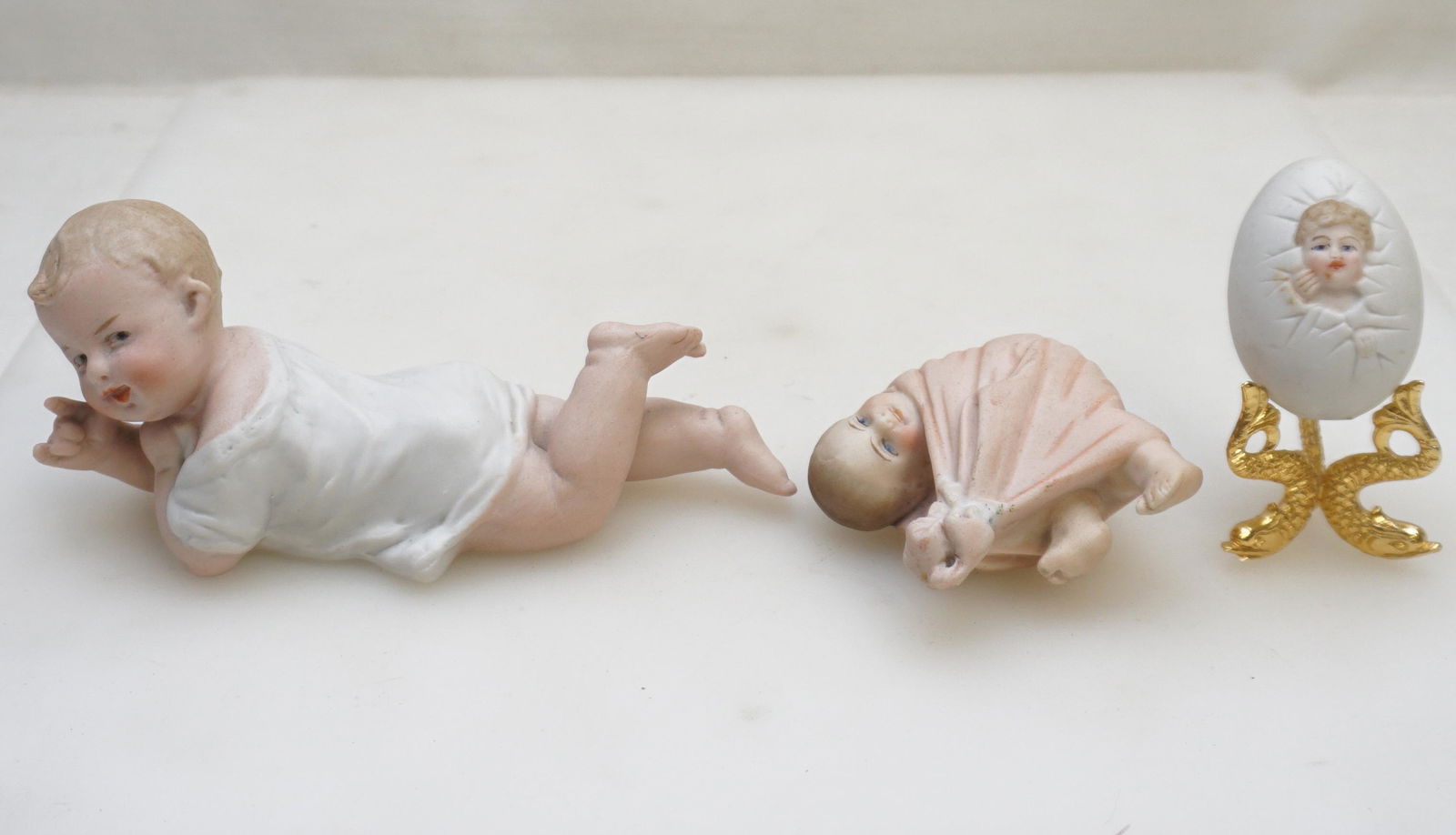 3 ANTIQUE HEUBACH PORCELAIN BABIES: All are Heubach Porcelain. 1- Rare bisque egg with a baby face and bare butt. It measures approximately 2 1/2" in height. With gilt dolphin stand. 2- Bisque Baby Girl in Pink Blanket 3". 3- Piano Baby