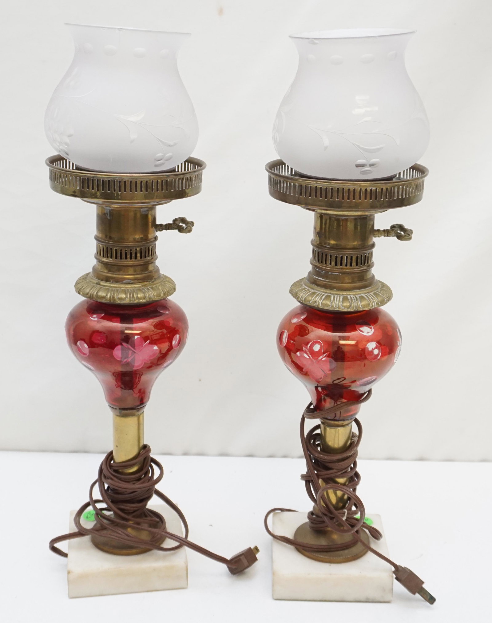 PAIR CRANBERRY CUT TO CLEAR LAMPS: Pair of Antique Electrified Cranberry cut-to-clear lamps with marble base and brass support column. Cut floral and leaf design with cut coin dots. Frosted globes with etched florals, both chipped. Lam