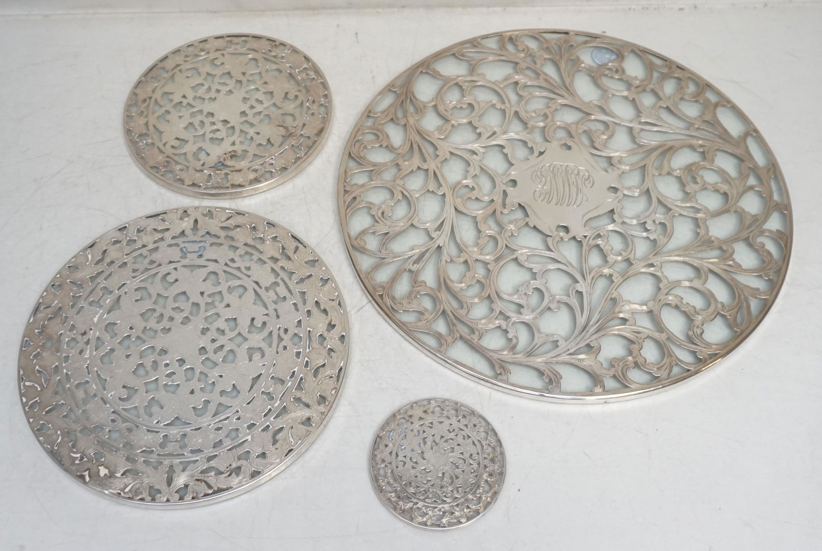 4 STERLING GLASS TRIVETS INCL 12": 4 American Ornate Sterling Silver and Glass Hot Plates The largest by Alvin measures 12 inches and is monogrammed. 1 Webster at 8". 1 Webster at 6". 1 Webster 3". Good condition. Total weight 5 pds 3