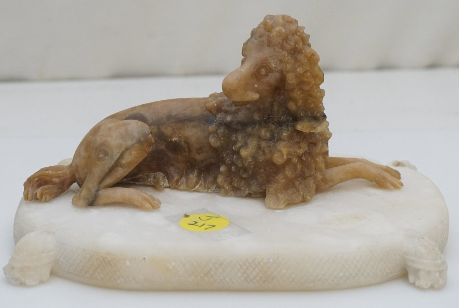 ALABASTER CARVING OF A POODLE: Brown alabaster recumbent poodle, mounted on a white alabaster pillow carved with a tassel at each corner; 6 3/4" x 4 1/2", 3 3/4"H; chips to tassels, one back tassel reattached, flea bites to poodle.