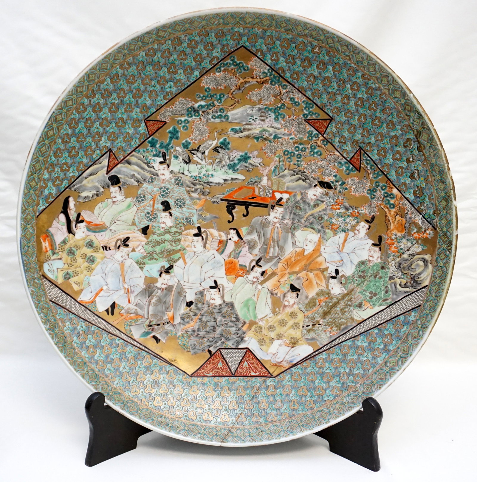 MONUMENTAL MEIJI PORCELAIN CHARGER: Heavily Enameled Meiji Deep Charger. Japanese, 19th century, center decorated with Samurai, Geisha, Pines. Raised on a circular foot with blue banding on base. Exterior with scrolling florals. Multipl