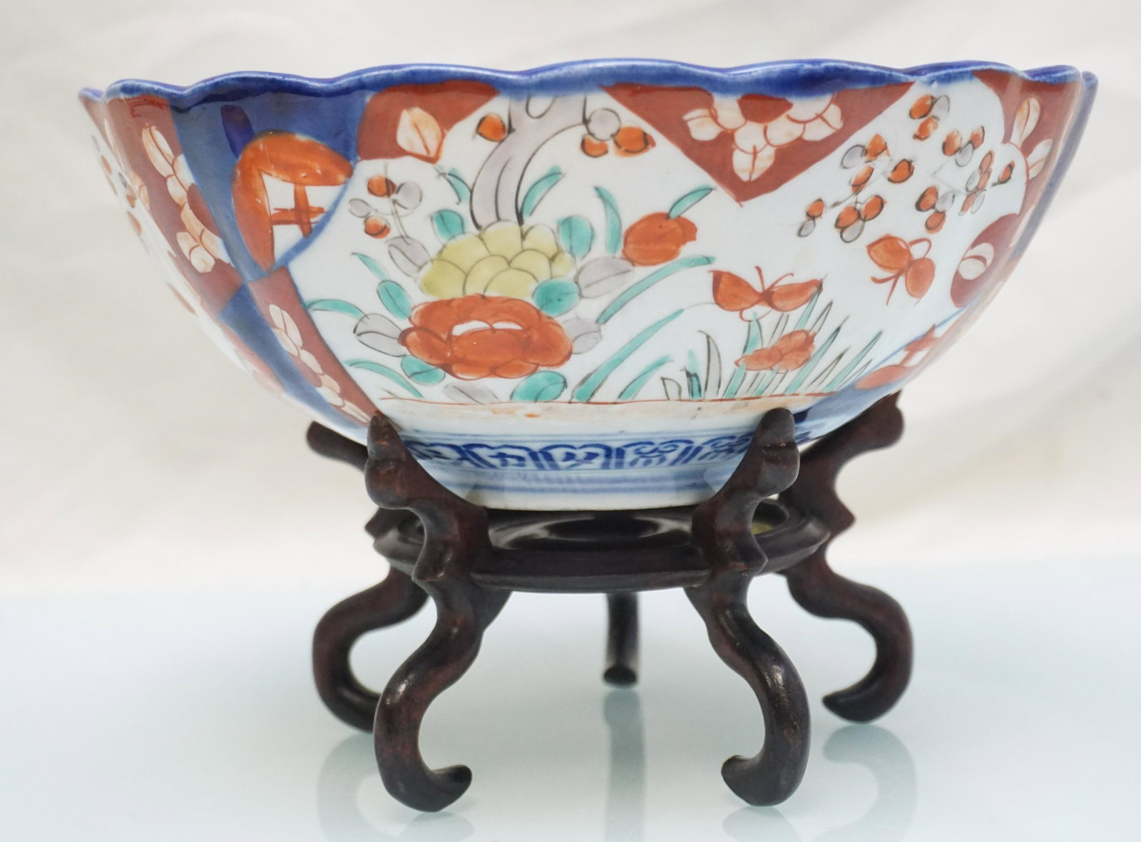JAPANESE IMARI BOWL WITH STAND: Antique Japanese Imari Bowl with Stand. Of lobed form, painted in underglaze blue with overglaze enamels, the inside decorated with a vase of flowerswithin a circular panel, and three panels with gard
