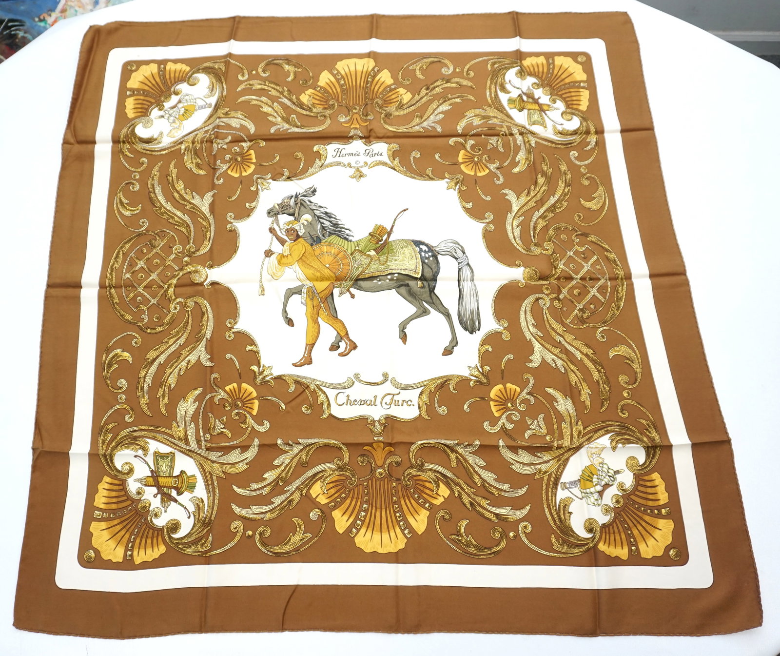 HERMES PARIS "CHEVAL TURC" SILK SCARF: HERMES PARIS silk "Cheval Turc" scarf. The scarf is designed with a white central motif displaying a horse being led by an African character. The scarf is accented with gold scroll work and a rich bro