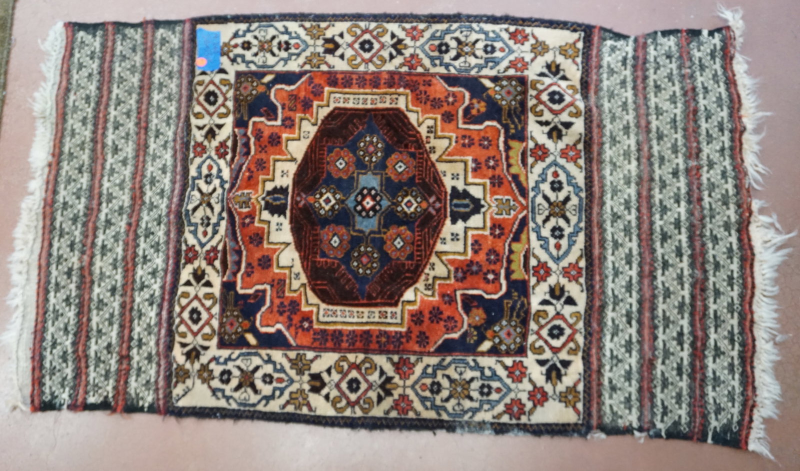 HAND WOVEN PERSIAN RUG: Hand made Persian rug, which perhaps could be transformed into a decoratibe pillow with the outter textule. The piece featyures a vibrant deep red center with cascading tones of tan, copper and blues.