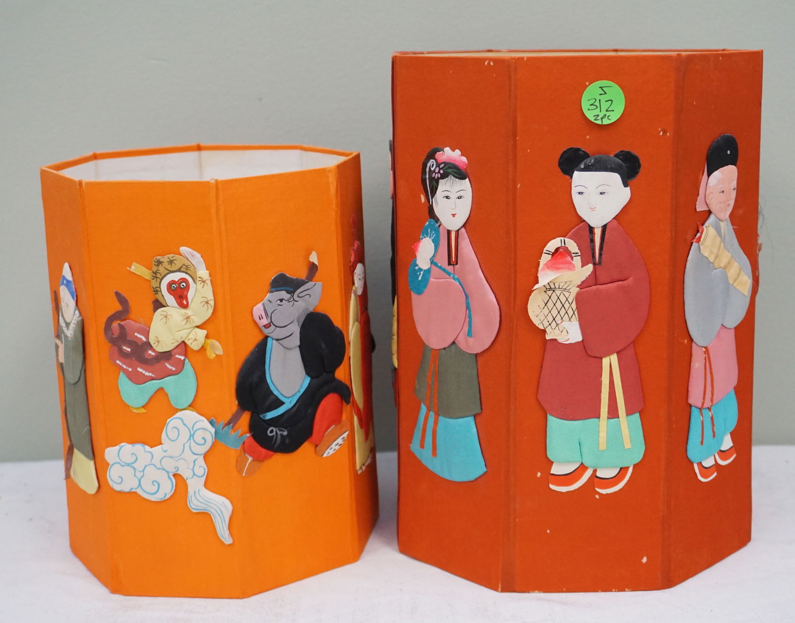 2 OCTAGONAL CHINESE SILK IMMORTALS BOXES: This set includes two decorative boxes, with the largest measuring 10" in height. Each box displays vibrant and colorful characters on an orange backdrop, suggesting a narrative or folklore significan