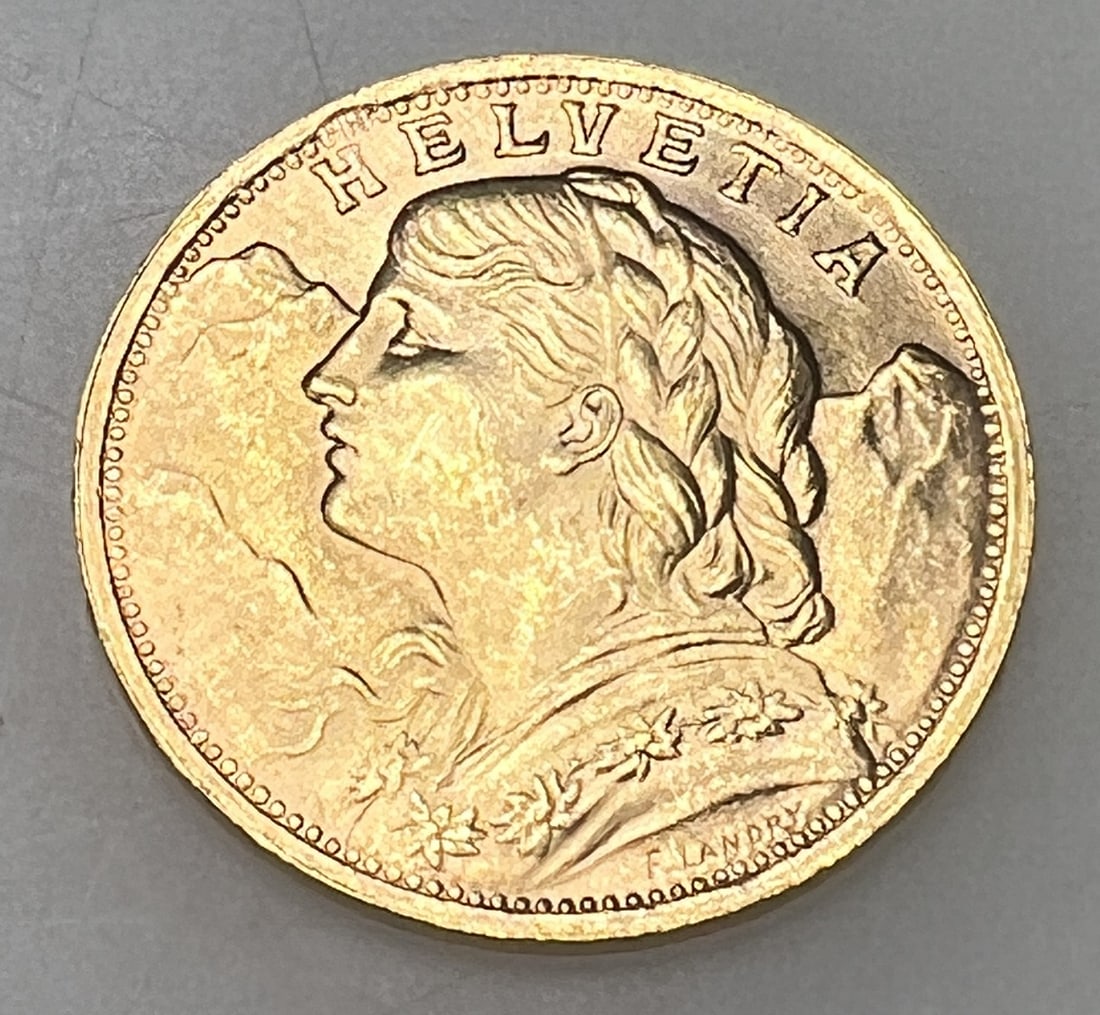 1935-LB SWISS HELVETIA 20 FRANC: Featuring the serene Swiss Miss, Vreneli, with the Alps softly detailed in the background, this 1935-LB Swiss Helvetia 20 Franc gold coin is a testament to Swiss craftsmanship. The reverse bears the S