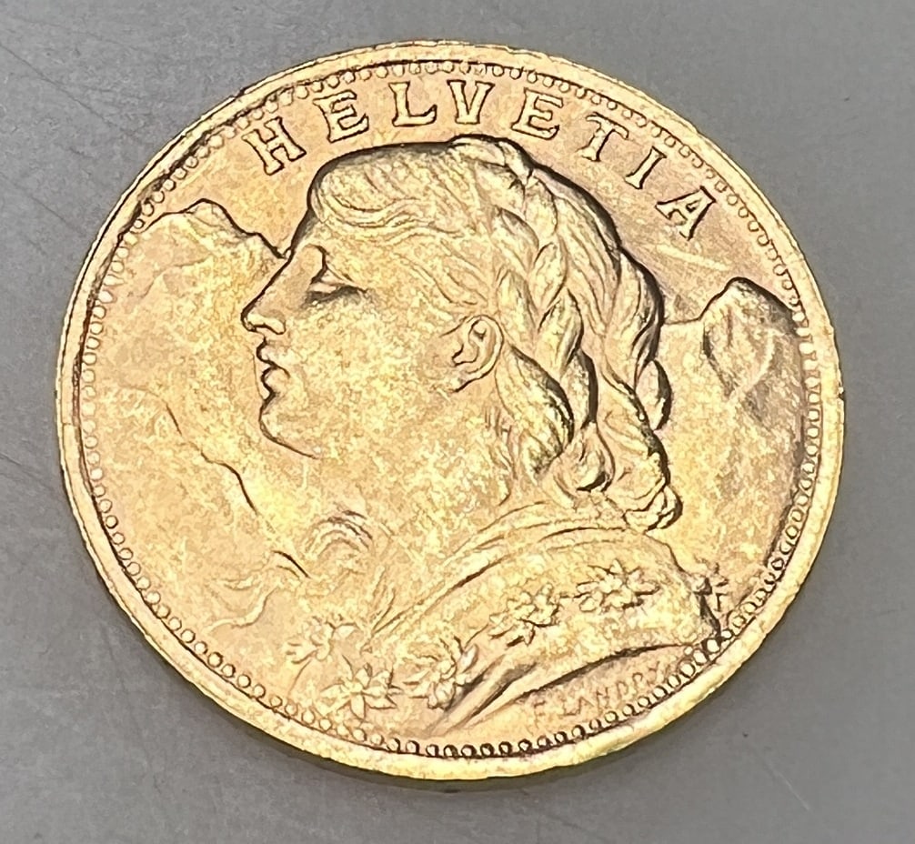 1935-LB SWISS HELVETIA 20 FRANC: Featuring the serene Swiss Miss, Vreneli, with the Alps softly detailed in the background, this 1935-LB Swiss Helvetia 20 Franc gold coin is a testament to Swiss craftsmanship. The reverse bears the S