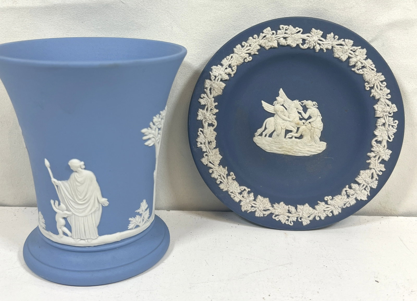 WEDGWOOD CUP AND PLATE: Two-piece Wedgwood set featuring a small blue cup and a matching plate. Both pieces are adorned with the classic white bas-relief design, showcasing intricate detail and craftsmanship. Cup measures ap