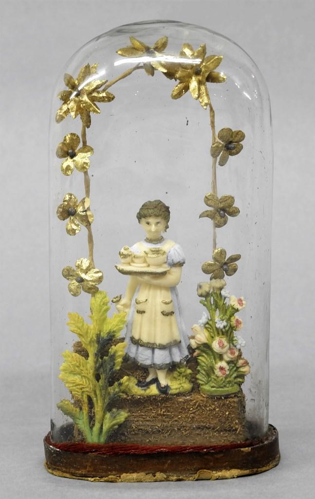 Glass Dome Diorama: Antique glass oval dome on a painted cardboard base contained a composition girl holding a tea set flanked by composition flowers and bush, surrounded by a ribbon of paper flowers and vine, 7.5"H, 4"W