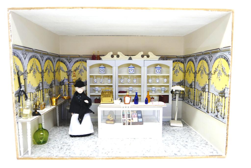 Small Furnished Pharmacy Room Box Dollhouse: Small plywood room box with paint and wallpaper covered walls, tiled floor, glass and wood counter contains a cash register, wood display box and artisan skull on book, three rear display cabinets hav