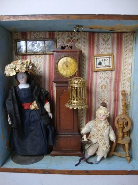 Framed Vignette with Ormolu Birdcage: A shadowbox with very old worm-eaten frame, containing a dressed china head doll, a bisque toddler, a vintage mahogany grandfather clock, a Tynietoy mirror, flax wheel and cello (both damaged) and a r