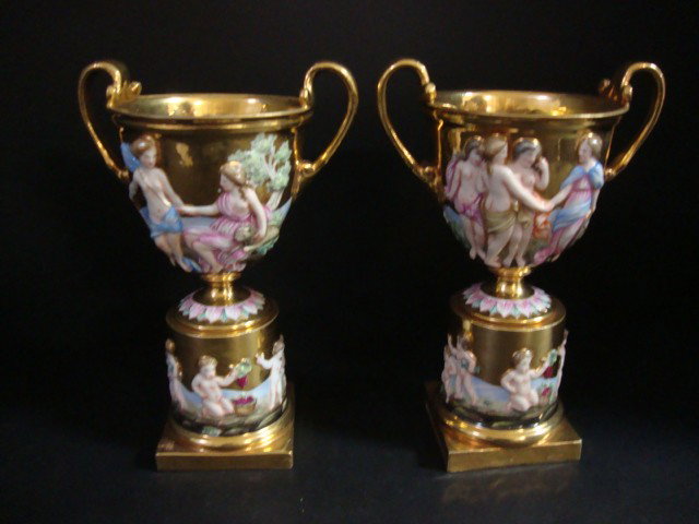Pair of Capodimonte Urns: Early Pair of gold gilt Urns on round pedestal set on square base, urns are profusely decorated w/raised nudes & cherubs, partial labels on bottoms 8 1/2" ( h)