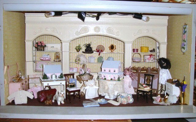 Contemporary Gift Shoppe Roombox: A roombox generously furnished with many appealing items including a pair of miniature dollhouses, many hats and decorated hatboxes, a handknit sweater and petipoint pillow, wrapped packages and femin