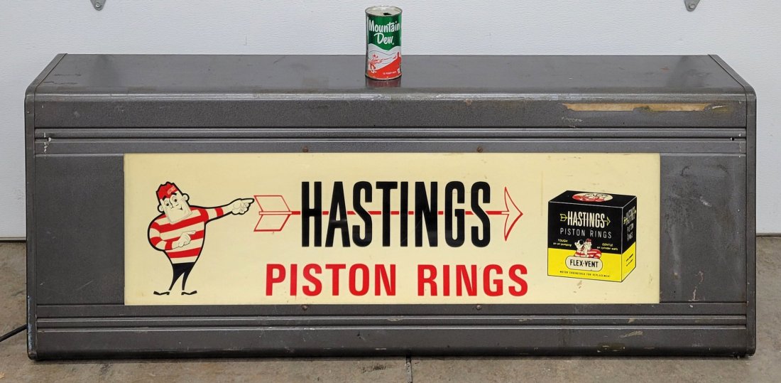 Hastings Piston Rings Light Up Auto Cabinet: 50.5" x 17" x 18"h metal cabinet with plastic sign showing bright colors. The cabinet shows normal wear with scuffs and scratches. The cabinet is electric with a light socket but no light bulb.