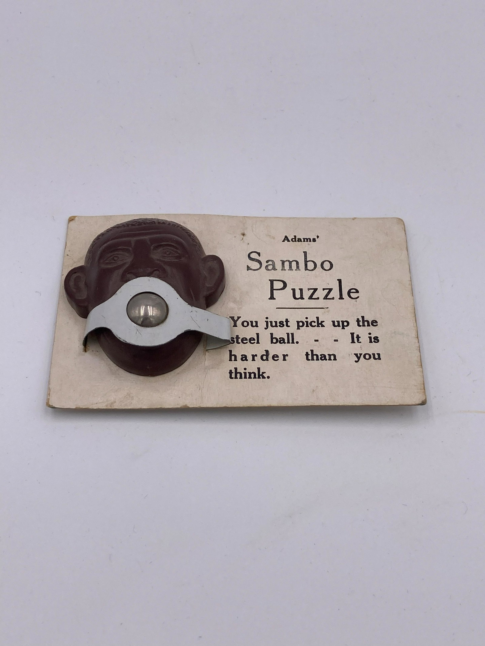 Adams' Sambo Puzzle Game: Adams' Sambo black americana puzzle game. Wear to corners of card and crease down center. Toy in good condition with metal ball and metal attachment. Measures 2.25" x 3.5". Please note that all items 