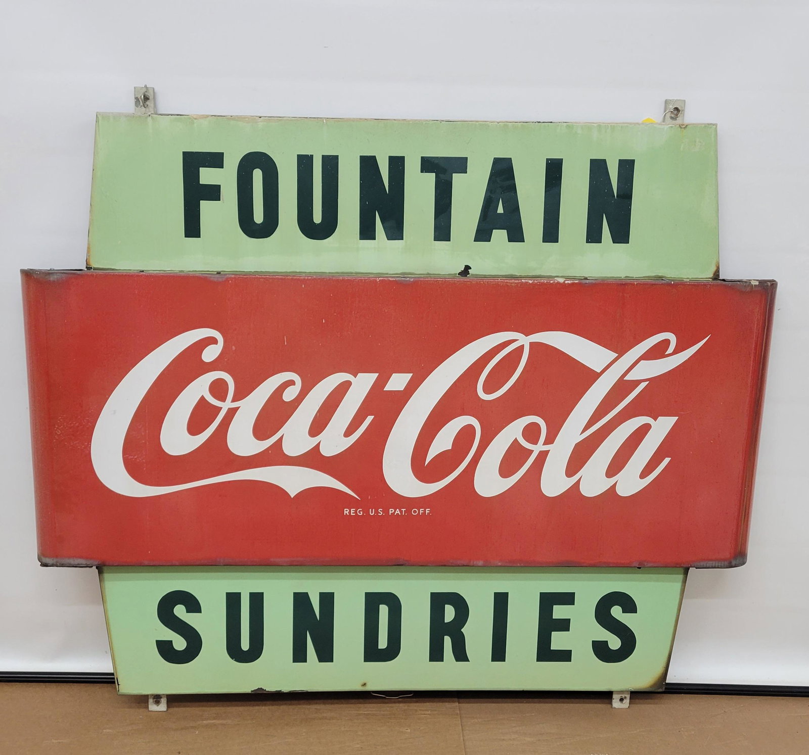 Coca Cola Fountain Sundries Single Sided Porcelain Sign: Coke. 60" x 52" Coca Cola sign that is made up of 3 separate SSP signs that are mounted on a metal bracket. The signs show normal wear with fading and stains. The top "Fountain" sign and bottom "Sundr