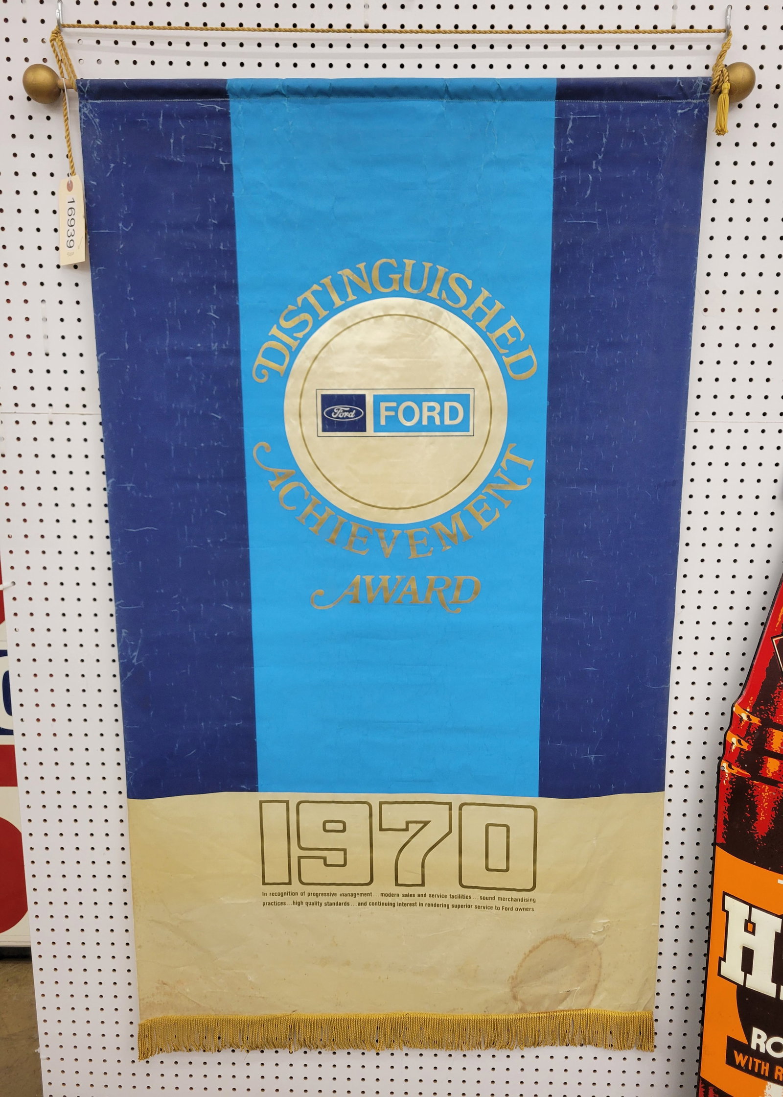 1970 Ford Advertising Banner: 35" x 60" 1970 Ford cloth advertising banner has light fold marks, stains at the bottom and several small holes. The bottom is accented with fringe trim. At the top, there is a wood dowel accented wit