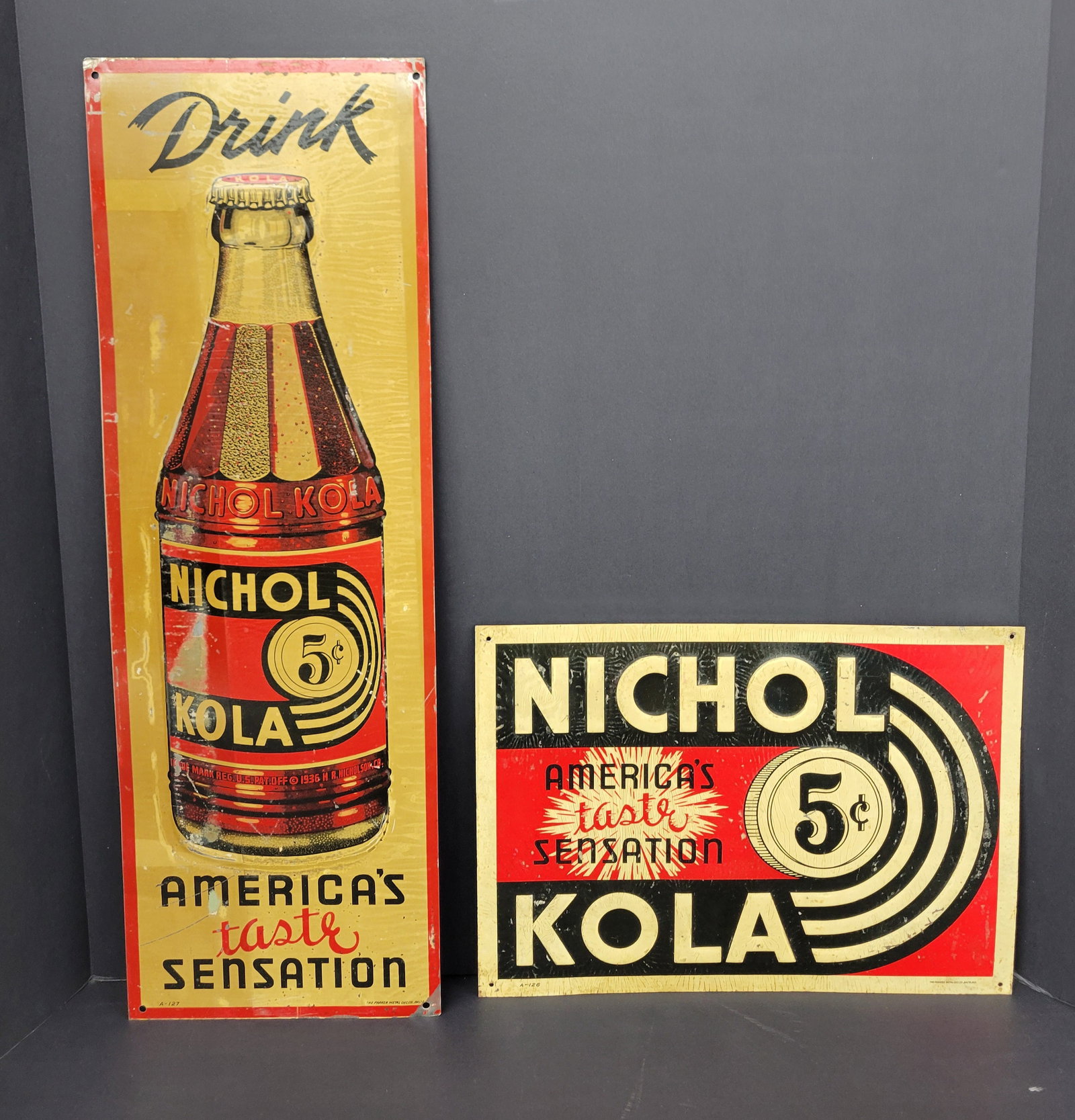 Nichol Kola Single Sided Tin Signs: The lot includes a 8" x 24" vertical SST sign that features an embossed bottle logo, which shows signs of discoloration, paint flakes, and scratches across its surface. The corners exhibit minor bends