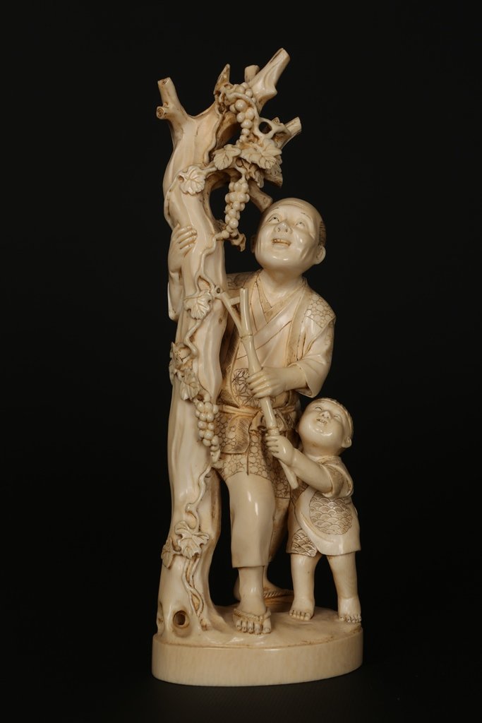 A Japanese ivory Okimono: A Japanese ivory Okimono, Meiji period, modelled as a farmer with a child at foot, standing beside a tree entwined with fruiting vines, signed plaque inset to base. 27cm