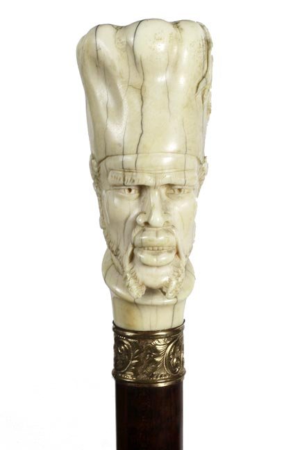 111. Ivory African King- Ca. 1910-A large carved: 111. Ivory African King- Ca. 1910-A large carved African king in head dress, ornate gold-filled collar, beautiful snake wood shaft and a metal ferrule. . H. 2 ¼” x 3 ¾”, O.L.-36” $800-$1,2