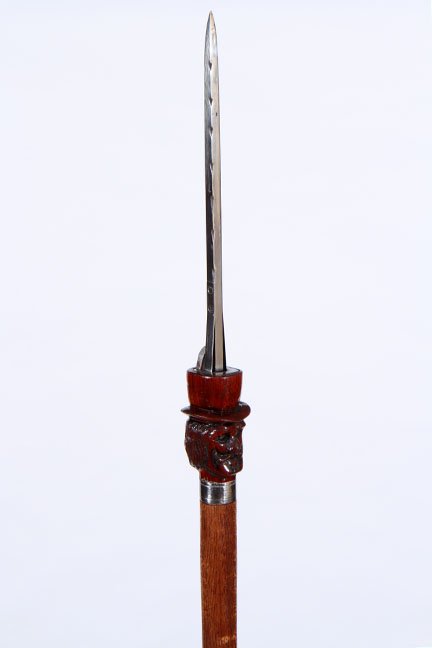 96. Flick Stick Cane- 20th Century- A carved wooden man: 96. Flick Stick Cane- 20th Century- A carved wooden man in a top hat with a flick stick mechanism atop, the four sided blade is 8” long and is in fine working condition, silver metal collar, oak sha
