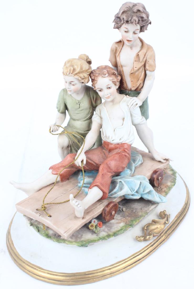 PORCELAIN FIGURINE THREE KIDS ON A SLED: PORCELAIN FIGURINE THREE KIDS ON A SLED Porcelain figurine of three kids playing on a wheeled sled. Capodimonte style. All setting on top of gold and white porcelain base. Marked "Benacch". Size: 9" x