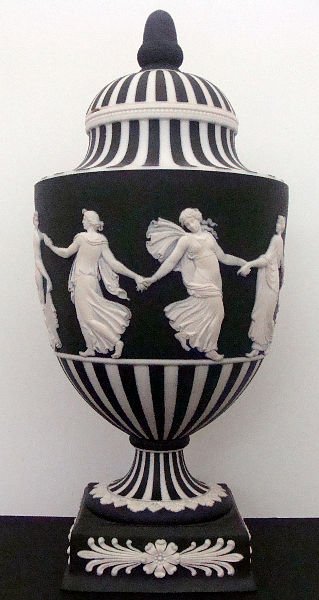 BLACK WEDGWOOD COVERED VASE: Black Wedgwood Covered Vase having raised white cameo design of women holding hands all around vase. Has a square base lid has two minor chips. Height 9 3/4" Diameter 4 1/2". SHIPPING & REQUIRED INSUR