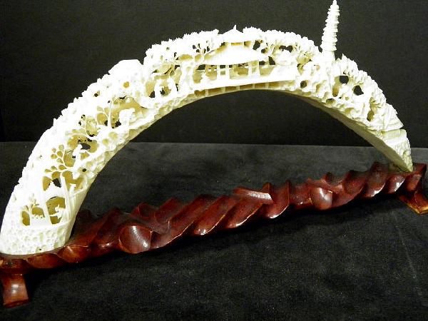 Heavily Carved Ivory Figural Tusk On Wood Base: Heavily carved ivory tusk. The whole displaying a village scene with temples, pagoda and figures walking and on horseback, trees and a boat in the foreground. Measures14 1/2" wide and 6 3/4" high. Shi