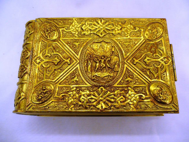 Dore Bronze Figural Book/Box: Dore bronze covered box. This box has the shape of a book and is decorated with raised figures and foliate scrolls throughout. The box is footed and has a black velvet interior. It measures 7 inches x