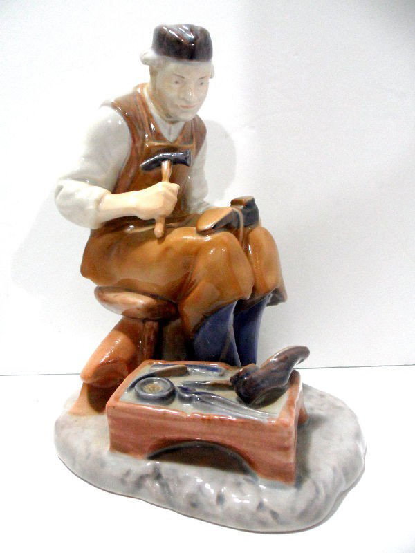 Bing & Grondahl (B&G) Figure of a Cobbler: A Bing & Grondahl (B&G) figure of a cobbler in glazed porcelain. The figure is sitting with a shoe on his lap and a work table next to him. The Bing & Grondahl stamp is glazed on the bottom. No: 2223,